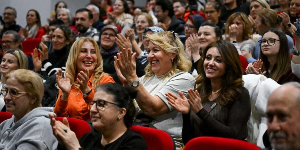 Kapalı bir salonda kırmızı koltuklarda oturan ve gülümseyerek alkışlayan kalabalık bir izleyici grubu.