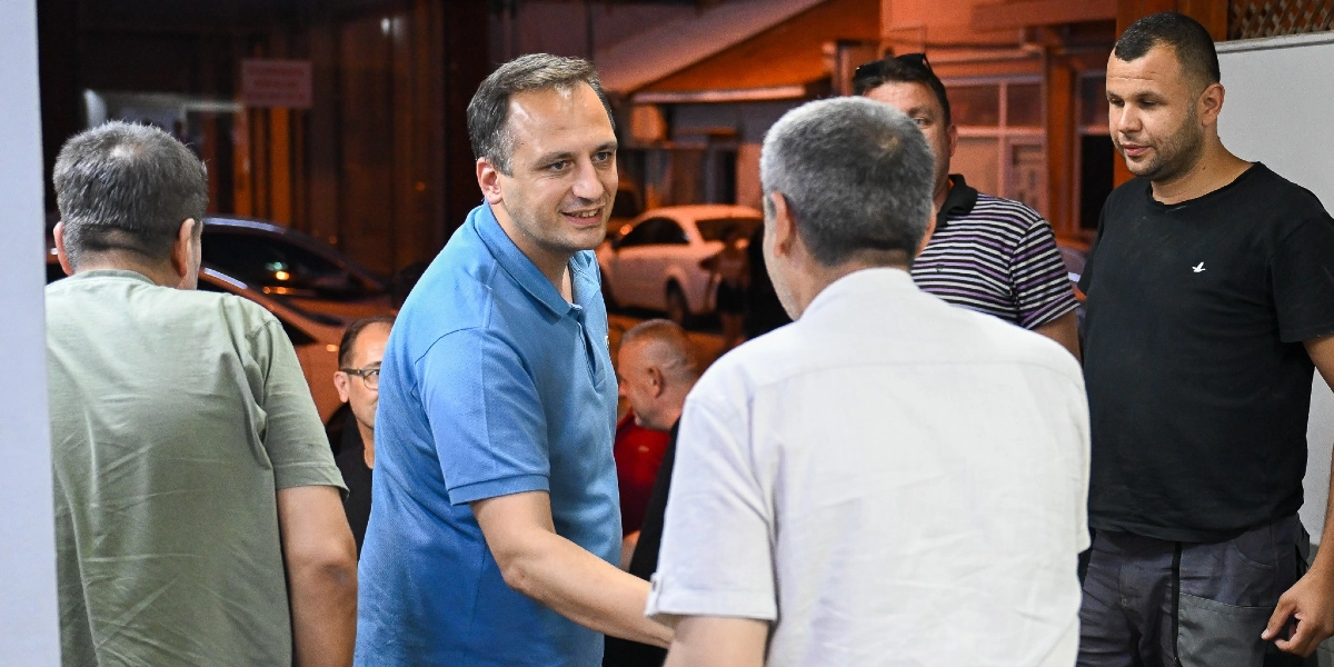 Mavi polo yaka tişört giyen bir erkek, akşam saatlerinde bir dükkanın önünde esnafla tokalaşarak sohbet ediyor. Çevresinde farklı yaşlarda birkaç erkek daha bulunuyor ve ortamda park halindeki araçlar görülüyor.