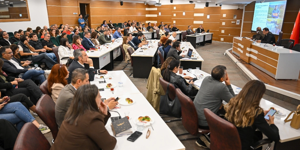 Modern bir konferans salonunda, U düzeninde yerleştirilmiş masalarda oturan kalabalık bir katılımcı grubu, ön taraftaki dev ekranda sunum yapan konuşmacıyı dinliyor. Masaların üzerinde çay bardakları ve küçük atıştırmalık tabakları bulunuyor. Salonun ön kısmında, kürsüde oturan yetkililer ve arkalarındaki ekranda tarımsal hizmetlerle ilgili görsellerin yer aldığı bir sunum ekranı görülüyor. Katılımcıların çoğu dikkatle dinlerken, bazıları not alıyor veya telefonlarıyla ilgileniyor.