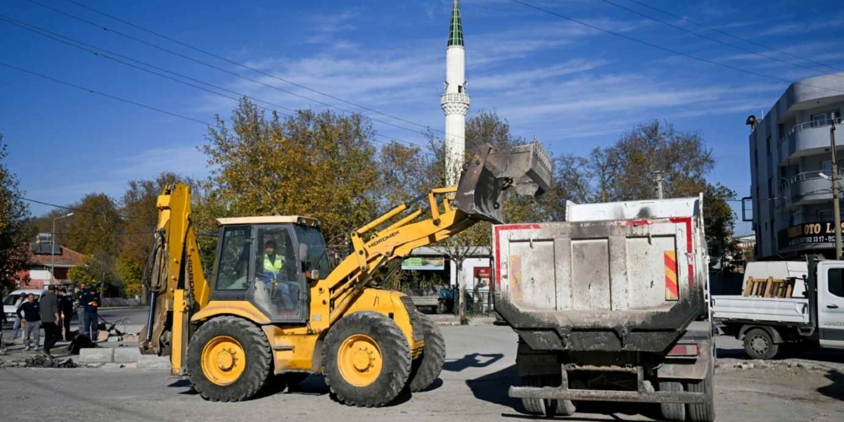 Bornova'da bir yol çalışması alanında, sarı renkli bir Hidromek iş makinesi kepçesindeki hafriyatı yanındaki kamyonun kasasına boşaltıyor. Çalışma alanının arka planında bir cami minaresi, ağaçlar ve sağ tarafta binalar görülüyor. İş makinesinin yanında ise birkaç belediye çalışanı bulunuyor.