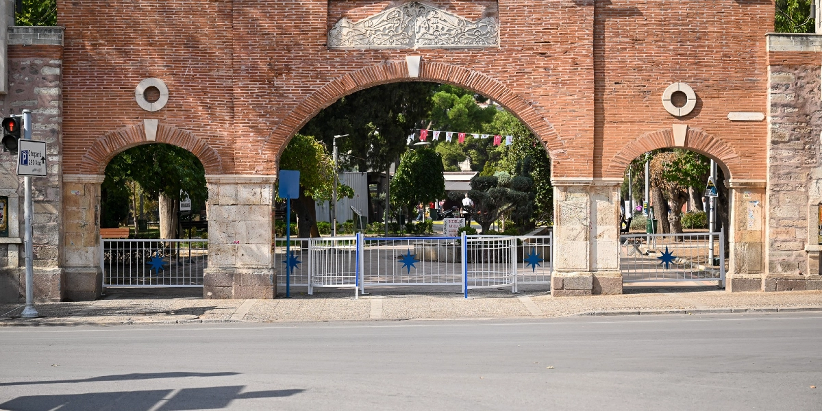 Bornova'daki tarihi bir park girişinin önünde, motosiklet girişini engellemek amacıyla yerleştirilmiş beyaz renkli metal bariyerler bulunuyor. Kemerli tuğla yapının giriş kısmını tamamen kapatan bu bariyerlerin üzerinde mavi yıldız süslemeleri yer alıyor. Parkın iç kısmında ağaçlar ve asılı Türk bayrakları görülürken, sol taraftaki direk üzerinde otopark yönlendirme tabelası dikkat çekiyor.