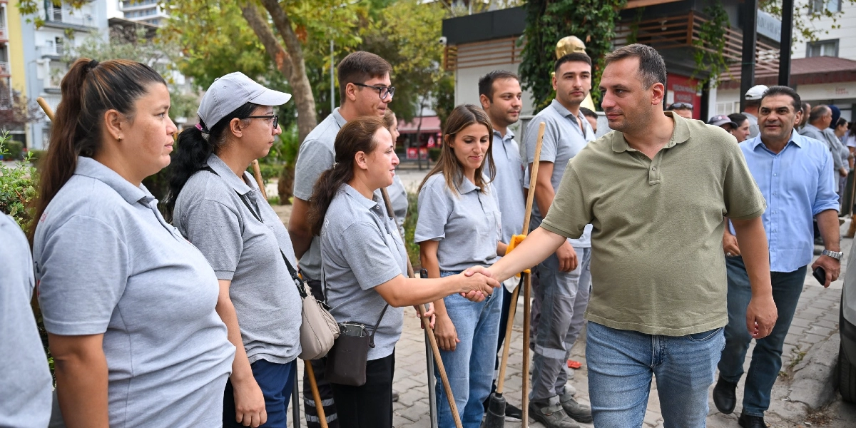 Açık havada, üzerinde gri tişörtler bulunan ve ellerinde temizlik süpürgeleri tutan bir grup belediye çalışanı ile yeşil polo yaka tişörtlü bir erkek tokalaşıyor. Grubun yanında açık mavi gömlekli bir başka erkek daha bulunuyor. Arka planda ağaçlar ve Bornova Belediyesi'ne ait olduğu anlaşılan bir yapı yer alıyor.
