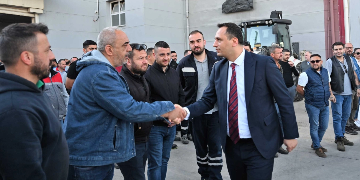 Lacivert takım elbiseli ve kırmızı çizgili kravatlı bir erkek, dış mekanda bir grup işçiyle tokalaşıyor. Çevresindeki çoğu gündelik kıyafetli olan kalabalık grup, bu anı izliyor. Arka planda gri bir bina duvarı ve bir iş makinesinin ön kısmı görünüyor.