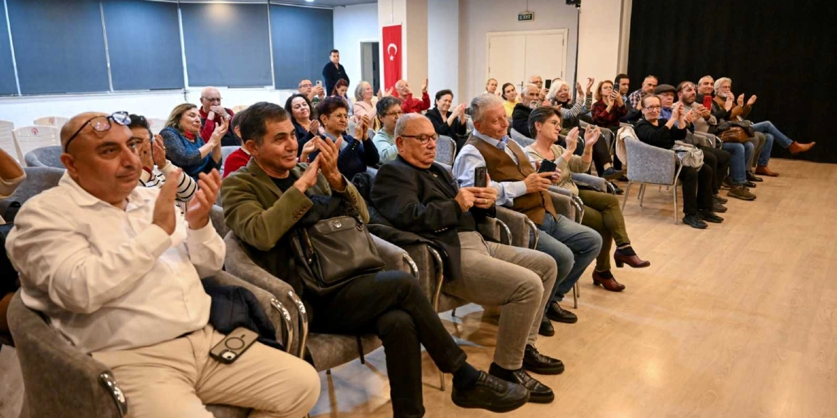 Kapalı bir salonda düzenlenen etkinlikte, gri koltuklarda oturan ve alkışlayan orta yaş ve üzeri izleyici grubu. Bazı katılımcılar cep telefonlarıyla fotoğraf çekerken, arka planda Türk bayrağı asılı duruyor.