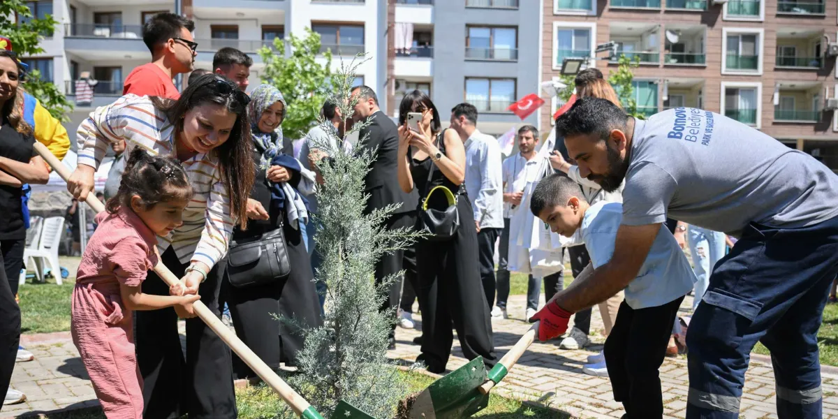 Bornova Belediyesi Park ve Bahçeler Müdürlüğü personeli, bir çocukla birlikte fidan dikimi yapıyor. Etrafta bu anı izleyen ve fotoğraflayan bir grup insan bulunuyor. Arka planda ise çok katlı konut binaları ve Türk bayrağı görülüyor.