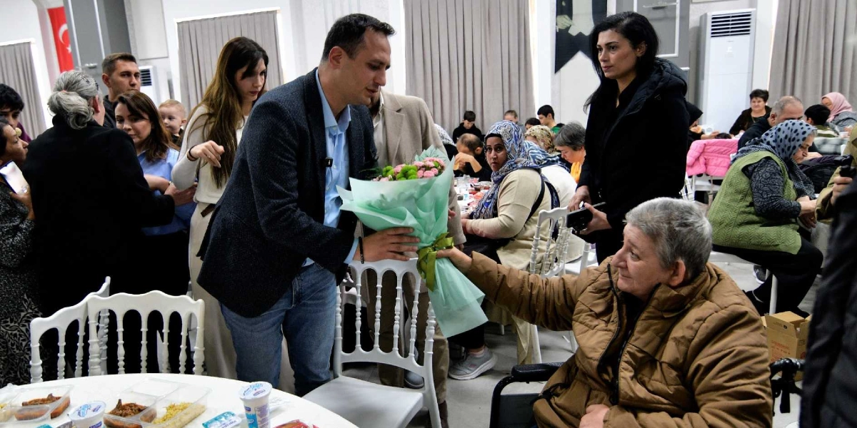 Bornova Belediye Başkanı Ömer Eşki, yılbaşı yemeğinde tekerlekli sandalyede oturan yaşlı bir vatandaşa çiçek takdim ediyor. Kapalı bir salonda düzenlenen yemekte, masalarda oturan çok sayıda Bornovalı vatandaş ve etkinlik görevlileri yer alıyor.