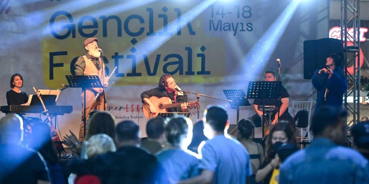Açık havada düzenlenen Gençlik Festivali sahnesinde, arkada 14-18 Mayıs yazılı dev bir afişin önünde müzik grubu performans sergiliyor. Grubun solisti, sakallı ve kasketli bir erkek ayakta şarkı söylüyor; yanında gitar çalan bir erkek, klavye başında bir kadın ve sağ tarafta ayakta şarkı söyleyen başka bir kadın yer alıyor. Sahne ışıklarıyla aydınlatılan grubun önünde, konseri izleyen kalabalık bir izleyici kitlesi bulunuyor.