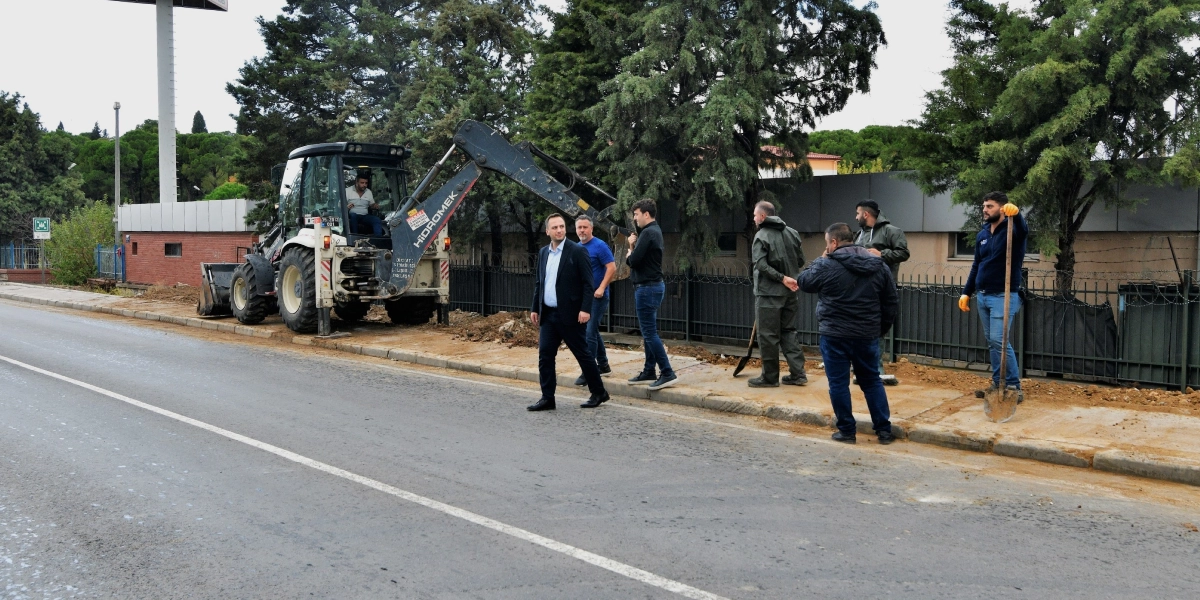 Koyu renk takım elbiseli bir yetkili, yol kenarındaki kaldırım çalışmasını incelemek üzere bir grup çalışanla birlikte şantiye alanında yürüyor. Arka planda Hidromek marka bir iş makinesi ve operatörü görülürken, diğer çalışanlar ellerinde küreklerle kaldırım düzenleme faaliyetine eşlik ediyor.