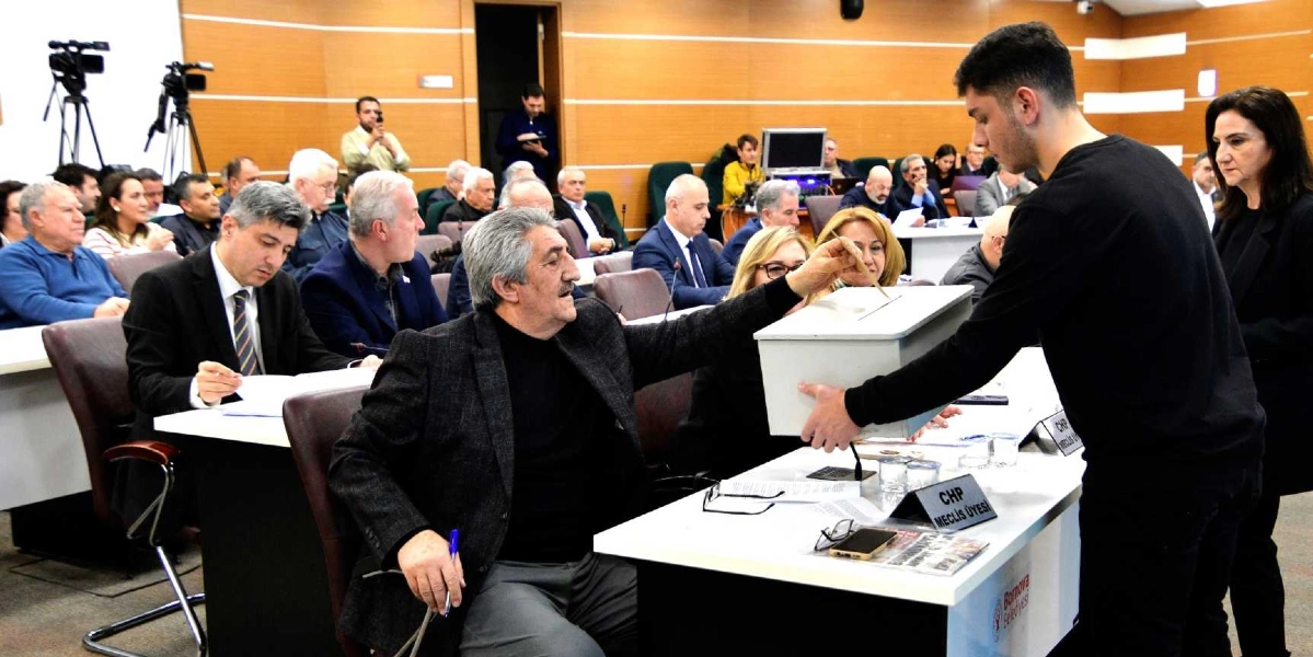 Bornova Belediyesi meclis salonunda gerçekleşen bir oylama sırasında, siyah kazaklı genç bir erkek elindeki oy pusulasını beyaz bir sandığa atıyor. Sandığın başında oturan gri saçlı ve ceketli bir meclis üyesi, sandığı tutarak gence yardımcı oluyor. Masanın üzerinde 