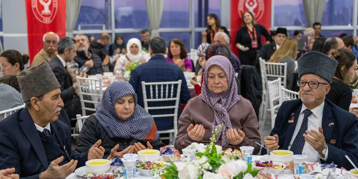 Modern bir salonda düzenlenen iftar yemeğinde, beyaz örtülü masaların etrafında oturan kalabalık bir grup insan görülüyor. Ön planda, geleneksel kalpak takan iki yaşlı adam ve başörtülü iki kadın, ellerini açmış dua ediyor. Masalarda çorba kâseleri, salatalar ve çeşitli yiyecekler bulunuyor. Arka planda Bornova Belediyesi logolu kırmızı flamalar asılı ve diğer davetliler yemeklerini yiyor.