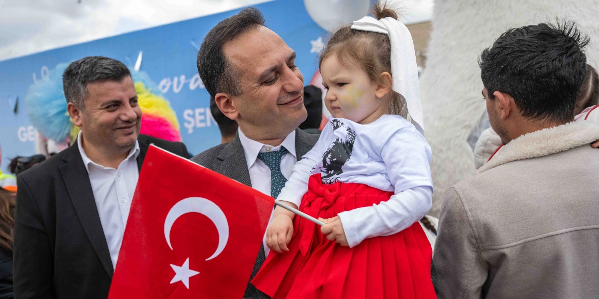Koyu renk takım elbiseli ve yeşil kravatlı bir adam, kucağında üzerinde Atatürk baskılı beyaz tişört ve kırmızı etek olan küçük bir kız çocuğunu tutuyor. Adamın yanında elinde Türk bayrağı tutan başka bir adam daha bulunuyor. Arka planda üzerinde 
