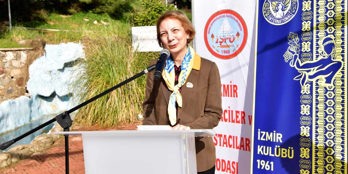 Açık havada, kürsü arkasında konuşma yapan orta yaşlı bir kadın. Kadın, kahverengi ceket ve sarı-mavi desenli bir fular giymiş. Kürsünün yanında üzerinde 