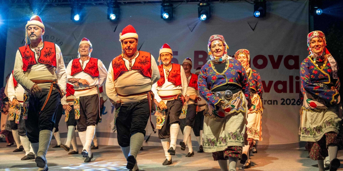 Sahne ışıkları altında geleneksel halk oyunları kıyafetleri giymiş bir grup dansçı, ellerinde tuttukları kaşıklarla ritmik bir şekilde dans ediyor. Erkek dansçılar beyaz gömlek, kırmızı yelek, koyu renk şalvar, beyaz tozluk ve kırmızı fes giyerken; kadın dansçılar çiçek desenli ceketler, işlemeli etekler ve başlıklar kullanıyor. Arka planda Bornova Festivali yazılı bir sahne dekoru bulunuyor.