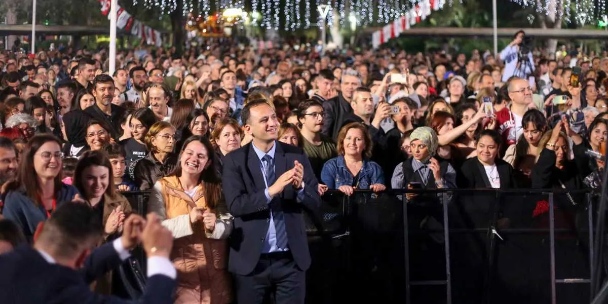 Gece vakti açık havada düzenlenen bir etkinlikte, bariyerlerin önünde toplanmış kalabalık bir grup insan görülüyor. Ön planda, lacivert takım elbiseli ve kravatlı bir erkek, gülümseyerek elleriyle alkış tutuyor. Yanında ve arkasında, farklı yaş gruplarından kadın ve erkeklerden oluşan, neşeli ve ilgili görünen büyük bir izleyici kitlesi yer alıyor. Arka planda ise ağaçların arasından sarkan ışıklandırmalar ve Türk bayrakları dikkat çekiyor.