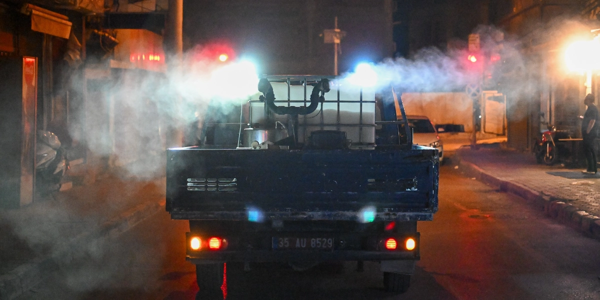 Gece vakti boş bir sokakta ilerleyen, arkasında büyük bir su tankı bulunan ve etrafa yoğun bir şekilde ilaçlama dumanı yayan mavi renkli bir kamyonet.