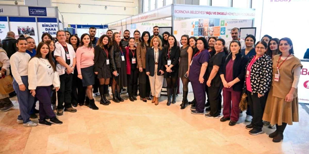 Bornova Belediyesi Yaşlı ve Engelli Hizmetleri Müdürlüğü standı önünde bir araya gelen, farklı kıyafetler giymiş ve yaka kartları takan kalabalık bir grup kadın ve erkek çalışan, gülümseyerek kameraya poz veriyor. Arka planda belediyenin hizmetlerini tanıtan afişler ve stantlar yer alıyor.