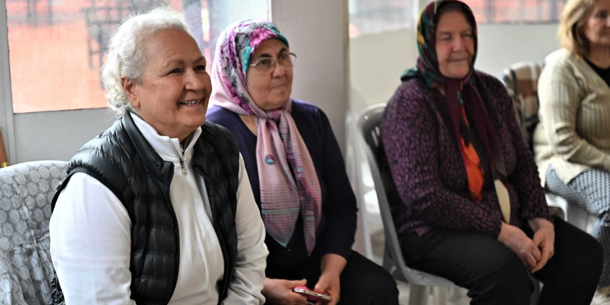 Yaratıcı drama etkinliğine katılan, yan yana sandalyelerde oturan ve gülümseyerek bir yöne bakan orta yaş üstü dört kadın.