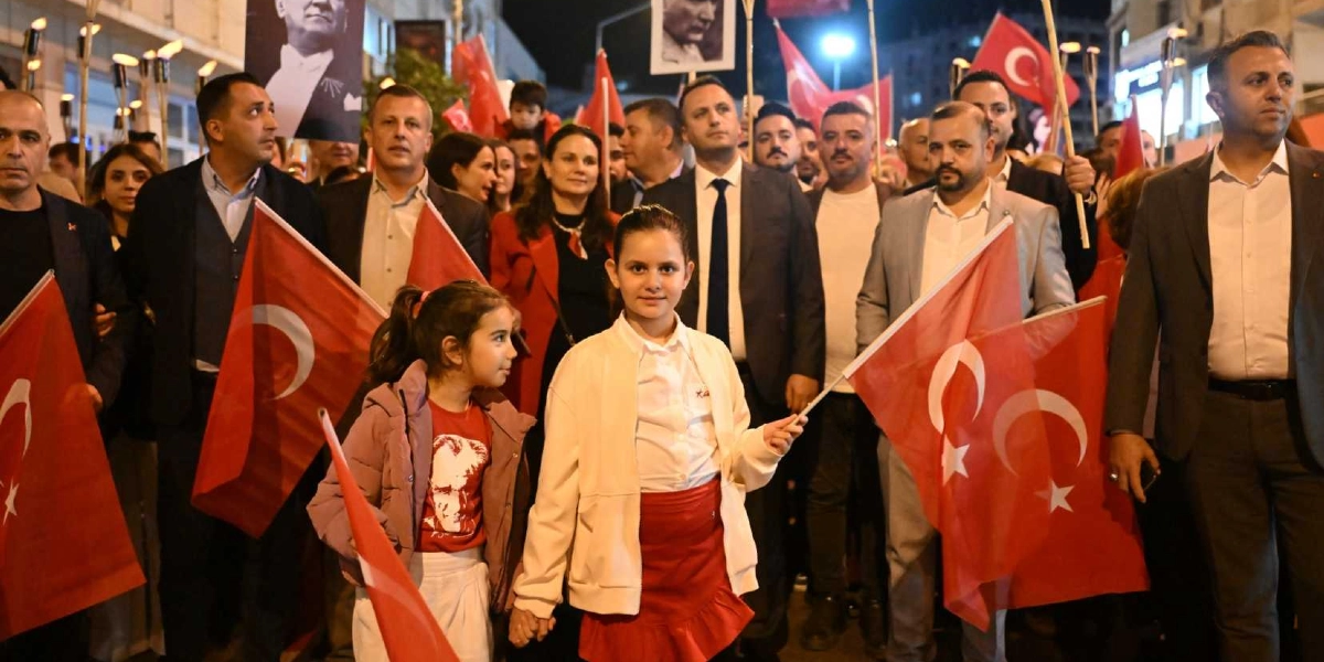 Gece vakti düzenlenen bir yürüyüşte ellerinde Türk bayrakları taşıyan kalabalık bir grup yer alıyor. Ön planda, ellerini tutan iki kız çocuğu bulunuyor; sol taraftaki çocuk üzerinde Atatürk baskılı kırmızı bir tişört ve pembe bir mont, sağ taraftaki çocuk ise beyaz bir ceket ve kırmızı bir etek giyiyor. Arka planda ise takım elbiseli yetişkinler ve ellerinde Atatürk posterleri taşıyan katılımcılar görülüyor.