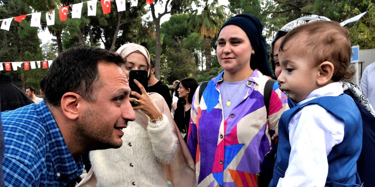 Mavi ekose gömlekli bir adam, kucağında bebek olan bir kadına ve yanındaki diğer kişilere gülümseyerek bakıyor. Kadınlardan biri elindeki telefonla fotoğraf çekerken, diğer kadın renkli desenli bir gömlek ve lacivert bir başörtüsü giyiyor. Arka planda ağaçlar ve Türk bayraklarıyla süslenmiş bir dış mekan alanı görülüyor.