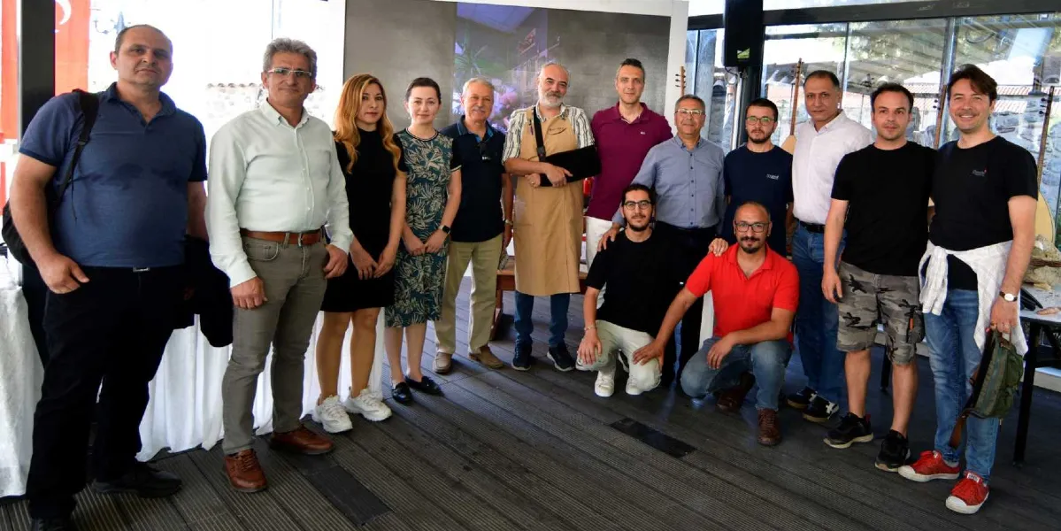 Aydınlık bir iç mekanda, yan yana duran ve bir kısmı çömelmiş olan on beş kişilik bir grup insan. Gruptakilerin çoğu gündelik kıyafetler giyerken, ortada duran bir kişi üzerinde bej rengi bir önlükle yer alıyor ve kolunda askı bulunuyor. Grubun arkasında, üzerinde müzik aletlerinin görsellerinin olduğu büyük bir pano ve cam bölmelerin ardında dış mekan manzarası görülüyor. İnsanlar gülümseyerek kameraya bakıyor.