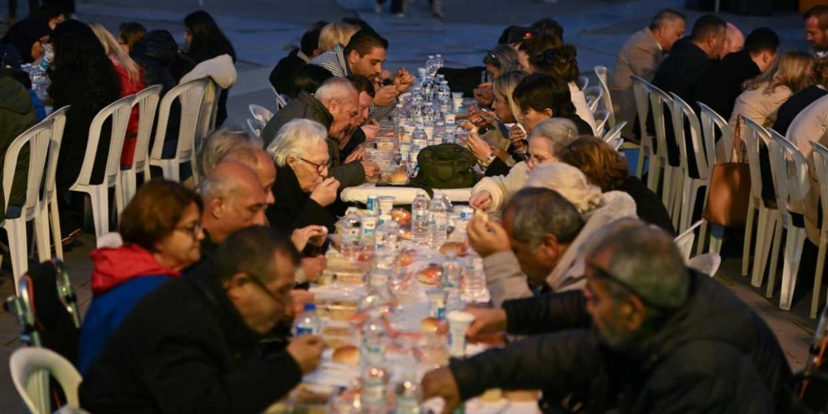 Açık havada, uzun bir masanın etrafında yan yana oturmuş çok sayıda kişinin katıldığı bir iftar yemeği. Beyaz plastik sandalyelerde oturan insanlar, önlerindeki masada bulunan yemek paketleri ve su şişeleri eşliğinde oruçlarını açıyor. Akşam karanlığında gerçekleşen buluşmada, farklı yaş gruplarından insanlar yemek yiyor ve sohbet ediyor.