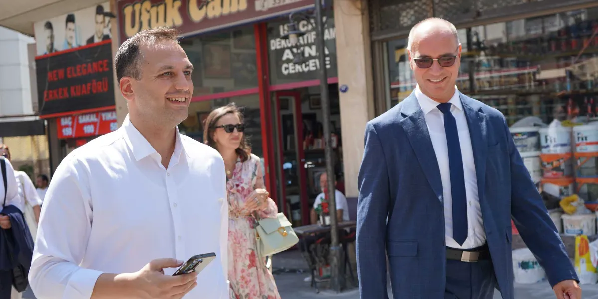 Beyaz gömlekli bir erkek, yanında lacivert takım elbiseli ve güneş gözlüklü bir başka erkekle birlikte dışarıda yürürken gülümsüyor. Arka planda, üzerinde 