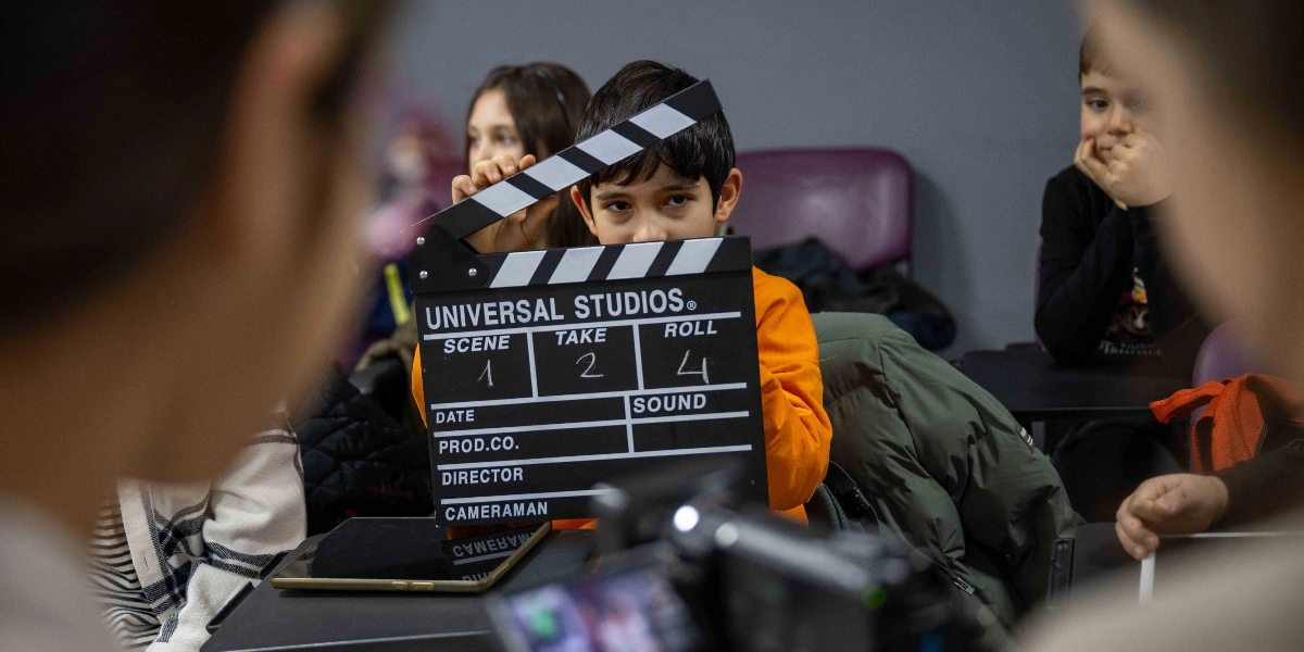 Turuncu tişörtlü bir çocuk, elinde tuttuğu Universal Studios yazılı film klaketini kapatırken kameraya bakıyor. Sınıf ortamında çekilen bu karede, çocuğun arkasında ve yanında diğer çocuklar dikkatle izliyor, ön planda ise bulanık bir kamera ekipmanı yer alıyor. Klaketin üzerinde sahne 1, çekim 2 ve rulo 4 bilgileri yazılı.
