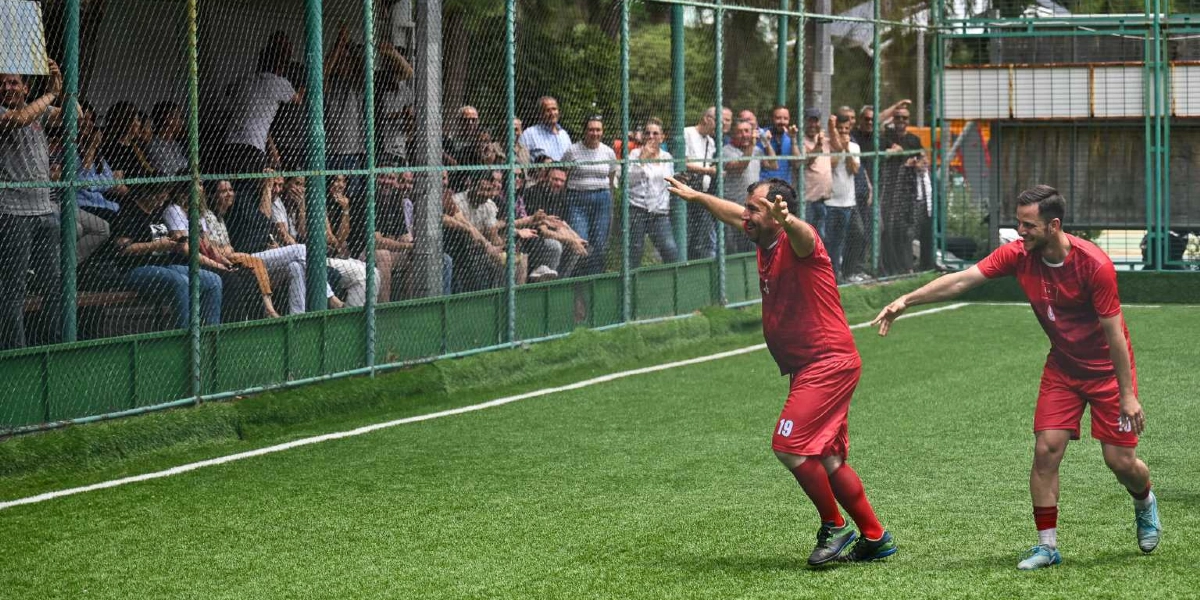Halı sahada kırmızı formalar giymiş iki futbolcu, attıkları golün ardından sevinçlerini paylaşıyor. 19 numaralı formayı giyen oyuncu kollarını iki yana açarak neşeyle koşarken, yanındaki takım arkadaşı ona eşlik ediyor. Tel örgülerle çevrili sahanın kenarında ise maçı izleyen ve alkışlayan bir grup seyirci bulunuyor.