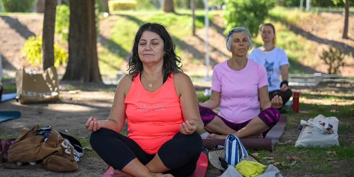 Ağaçlık bir park alanında, matlarının üzerinde bağdaş kurmuş ve gözleri kapalı şekilde meditasyon yapan üç kadın. Ön planda neon turuncu atlet giyen bir kadın, hemen arkasında pembe tişörtlü bir kadın ve onun arkasında beyaz tişörtlü bir kadın oturuyor. Kadınların etrafında yoga matları, bez çantalar ve kişisel eşyalar bulunuyor.