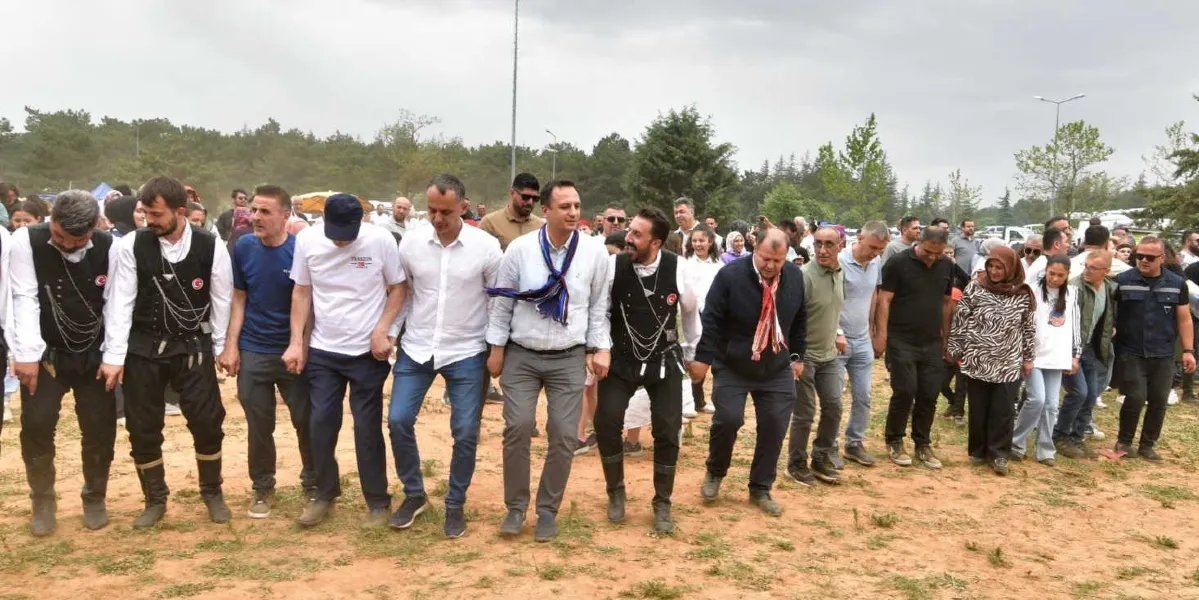 Açık havada, toprak bir alanda yan yana dizilmiş kalabalık bir grup insan, el ele tutuşarak Karadeniz yöresine ait horon oyununu oynuyor. Grubun ön sırasında geleneksel Karadeniz kıyafetleri giyenlerin yanı sıra, beyaz gömlekli ve boynunda mavi-beyaz bir fular bulunan Başkan Eşki, diğer katılımcılarla birlikte neşeli bir şekilde dansa eşlik ediyor. Arka planda ağaçlık bir alan ve bulutlu bir gökyüzü görülüyor.