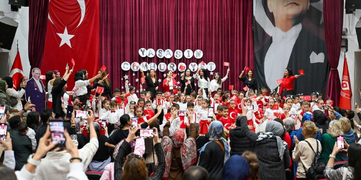Bir okul gösterisinde, bordo perdeli sahne önünde ellerinde küçük Türk bayrakları tutan anaokulu öğrencileri ve onlara eşlik eden öğretmenleri bulunuyor. Sahne arkasında 
