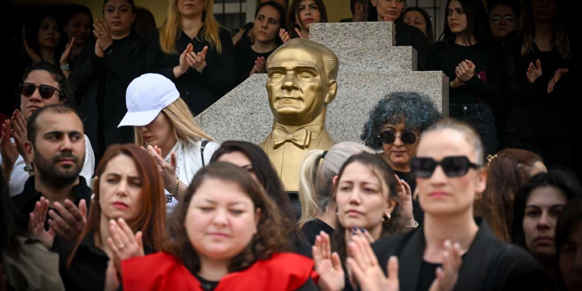 Mustafa Kemal Atatürk'ün altın rengi büstünün önünde toplanmış, çoğunluğu siyah kıyafetler giymiş bir grup insan, okullarda şiddeti protesto etmek amacıyla alkış tutuyor.