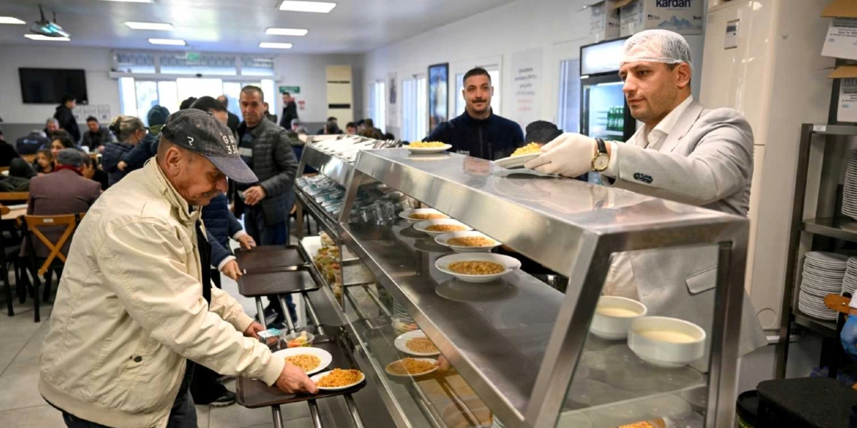 Bir yemekhanede, beyaz ceketli ve saç bonesi takmış bir görevli, eldivenli elleriyle servis tezgahının arkasından yaşlı bir adama tabak uzatıyor. Tezgahın üzerinde içinde yemek bulunan tabaklar sıralanmış durumda. Sıra bekleyen diğer insanlar ve arka planda masalarda oturan vatandaşlar, dayanışma ortamını yansıtıyor.