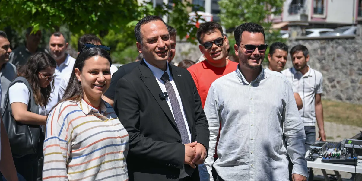 Açık havada düzenlenen bir etkinlikte, takım elbiseli bir yetkilinin yanında çizgili gömlekli bir kadın ve güneş gözlüklü bir erkek ile birlikte gülümsediği, arka planda ise diğer katılımcıların ve bir ses sisteminin yer aldığı grup fotoğrafı.