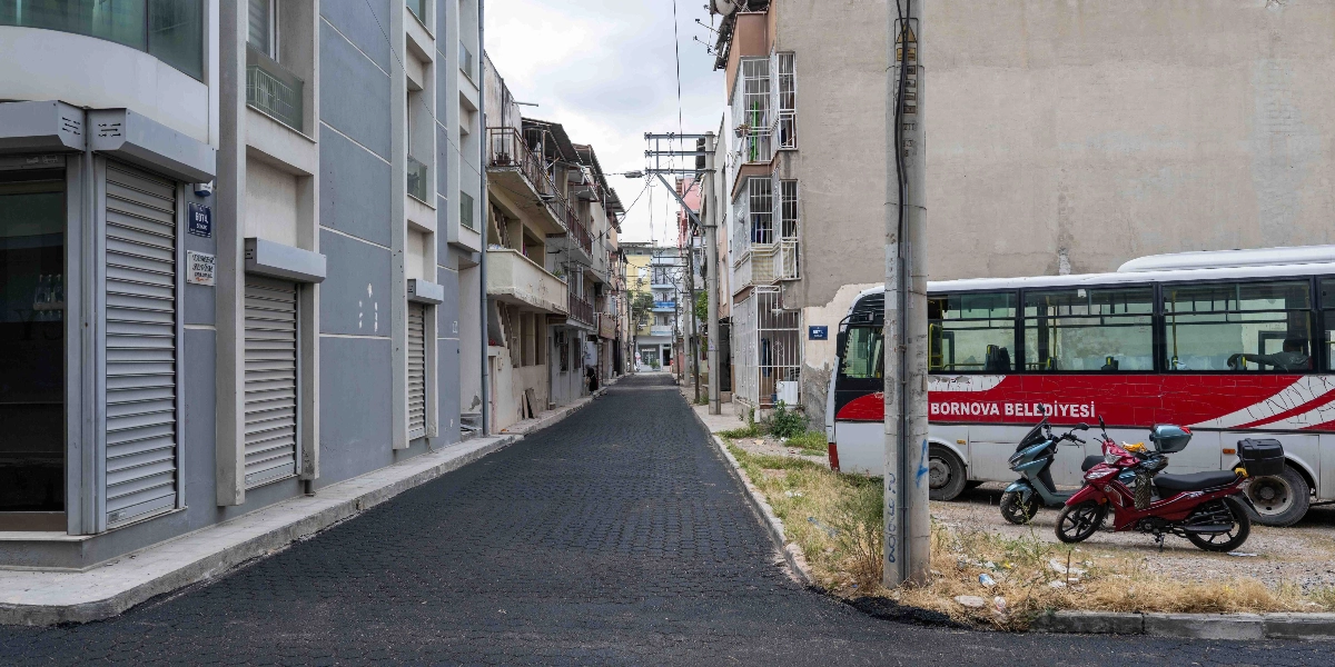 Bornova'da yeni asfaltlanmış, pürüzsüz ve koyu renkli bir sokak. Sokağın sağ tarafında Bornova Belediyesi'ne ait beyaz ve kırmızı renkli bir otobüs ile park halinde iki motosiklet bulunuyor. Sokağın her iki yanında ise çok katlı apartmanlar ve dükkanlar yer alıyor.