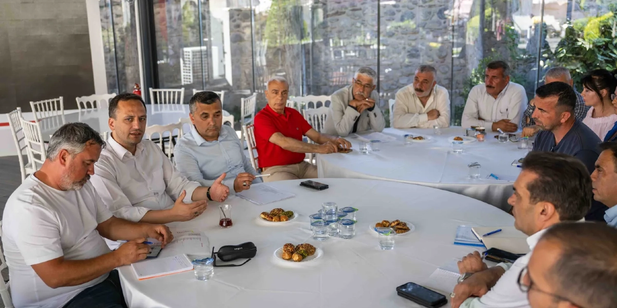 Aydınlık ve modern bir toplantı salonunda, beyaz örtülü büyük yuvarlak bir masa etrafında toplanmış, aralarında Bornova yerel yöneticilerinin ve muhtarların bulunduğu bir grup erkek ve kadın, sorunları görüşmek üzere bir araya gelmiş. Katılımcılar masada otururken bir yandan notlar alıyor, bir yandan da karşılıklı fikir alışverişinde bulunuyor. Masanın üzerinde küçük tabaklarda kurabiyeler, bardak suları ve çaylar yer alıyor. Arka planda cam duvarların ardında yeşil bir bahçe manzarası görülüyor.