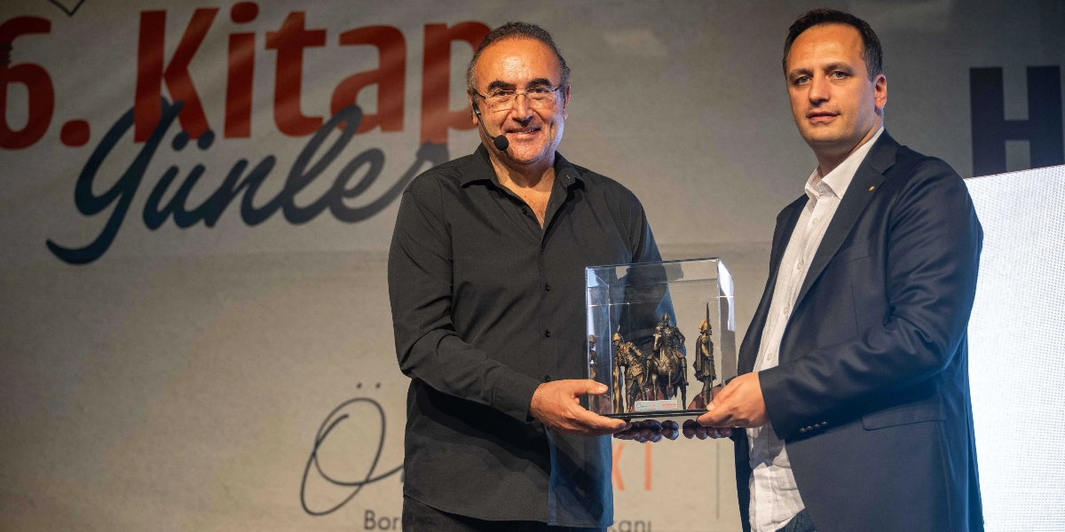 Siyah gömlekli bir erkek, yanında duran lacivert ceketli bir erkeğe, içinde atlı asker figürlerinin bulunduğu şeffaf bir plaket takdim ediyor. Arka planda 