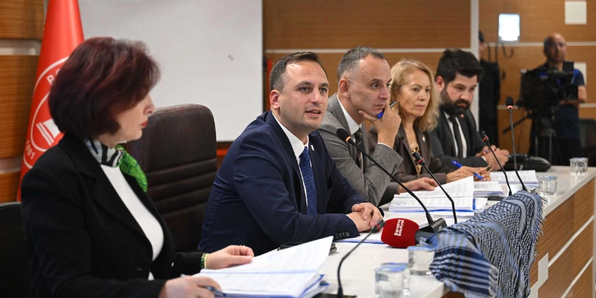 Bornova Belediye Meclisi toplantısında, masada oturan Bornova Belediye Başkanı Ömer Eşki ve meclis üyeleri konuşma yapıyor. Masanın üzerinde Bornova Belediyesi logolu bir mikrofon ve Filistin'i simgeleyen siyah beyaz puşi bulunuyor. Arka planda Bornova Belediyesi'nin kırmızı bayrağı ve toplantıyı takip eden bir kameraman yer alıyor.