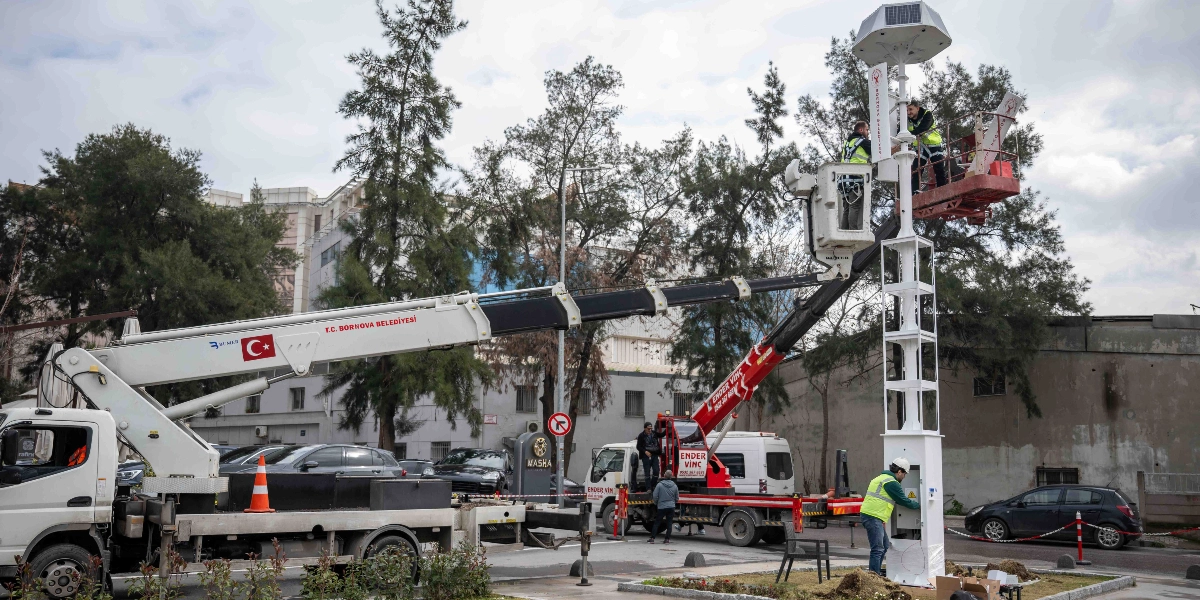 T.C. Bornova Belediyesi'ne ait bir vinç, üzerinde iki işçinin bulunduğu sepetiyle, güneş enerjisiyle çalışan yeni bir aydınlatma direğinin montajını yapıyor. Direğin tabanında bir başka işçi elektrik bağlantıları üzerinde çalışırken, çevrede park halindeki araçlar ve ağaçlar yer alıyor.