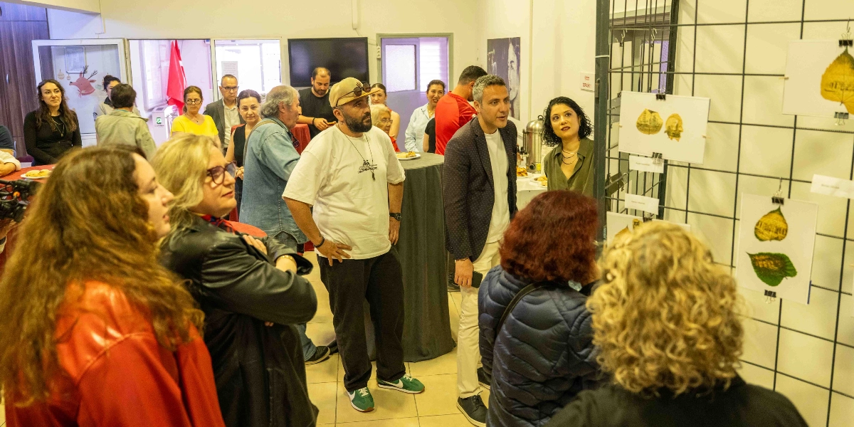 Kapalı bir mekanda düzenlenen sanat sergisinde, metal bir pano üzerine asılmış yaprak üzerine baskı çalışmalarını inceleyen bir grup insan yer alıyor. Ön planda sırtı dönük veya profilden görünen katılımcılar, sergilenen eserlere bakarken, orta alanda beyaz tişörtlü ve şapkalı bir erkek ile yanında lacivert ceketli bir başka erkek sohbet ediyor. Arka planda ise ayakta duran ve birbirleriyle etkileşimde olan çeşitli yaş gruplarından insanlar, kokteyl düzeninde bir etkinlik ortamında bulunuyor.