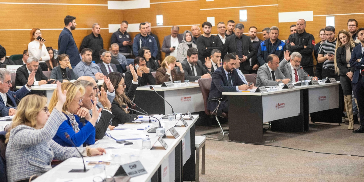 Bornova Belediyesi meclis salonunda düzenlenen toplantıda, meclis üyeleri masalarda otururken bir oylama sırasında ellerini kaldırarak oy kullanıyor. Salonun ön sıralarında yer alan meclis üyeleri, önlerinde mikrofonlar ve isimliklerle otururken, arka planda izleyiciler ve basın mensupları ayakta veya oturarak toplantıyı takip ediyor. Ahşap kaplama duvarlara sahip salonda, masaların üzerinde Bornova Belediyesi logoları yer alıyor.