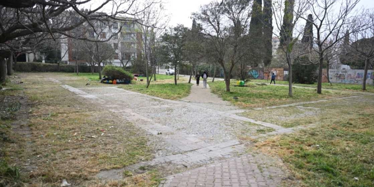 Ağaçlarla çevrili, yer yer çim ve toprak alanların bulunduğu parkın ortasında taş döşeli yürüyüş yolları yer alıyor. Parkın sol tarafında, ağaçların altında kediler için yapılmış küçük bir barınak ve renkli kedi evleri bulunuyor. Arka planda apartmanlar ve duvarlarında grafitiler olan bir yapı görülürken, parkın içinde yürüyen insanlar yer alıyor.