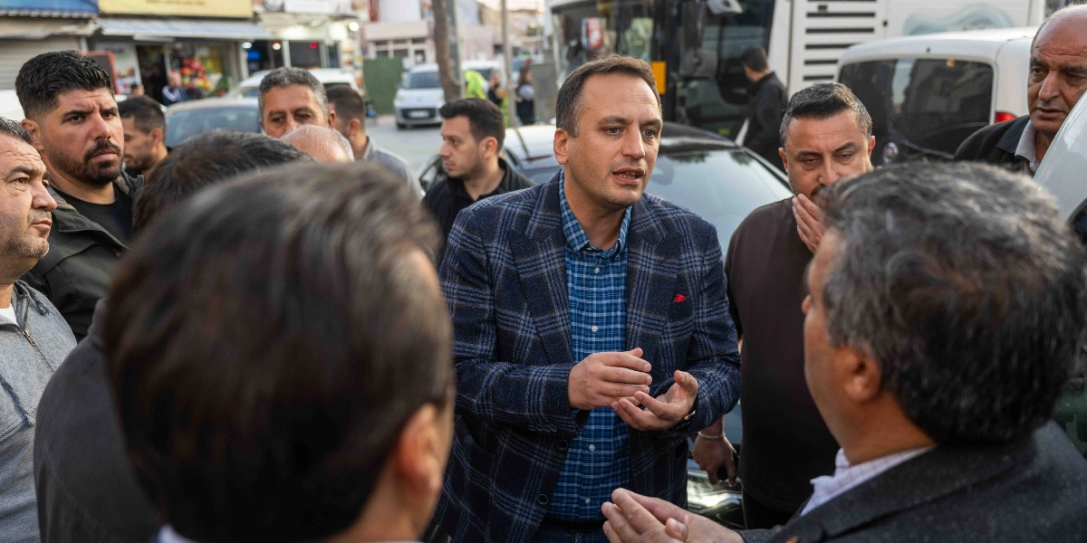Kareli ceket ve mavi gömlek giyen bir erkek, dış mekanda çevresini saran bir grup insanla konuşuyor ve elleriyle açıklayıcı bir jest yapıyor. Arka planda park halindeki araçlar ve hareketli bir sokak ortamı görülüyor.