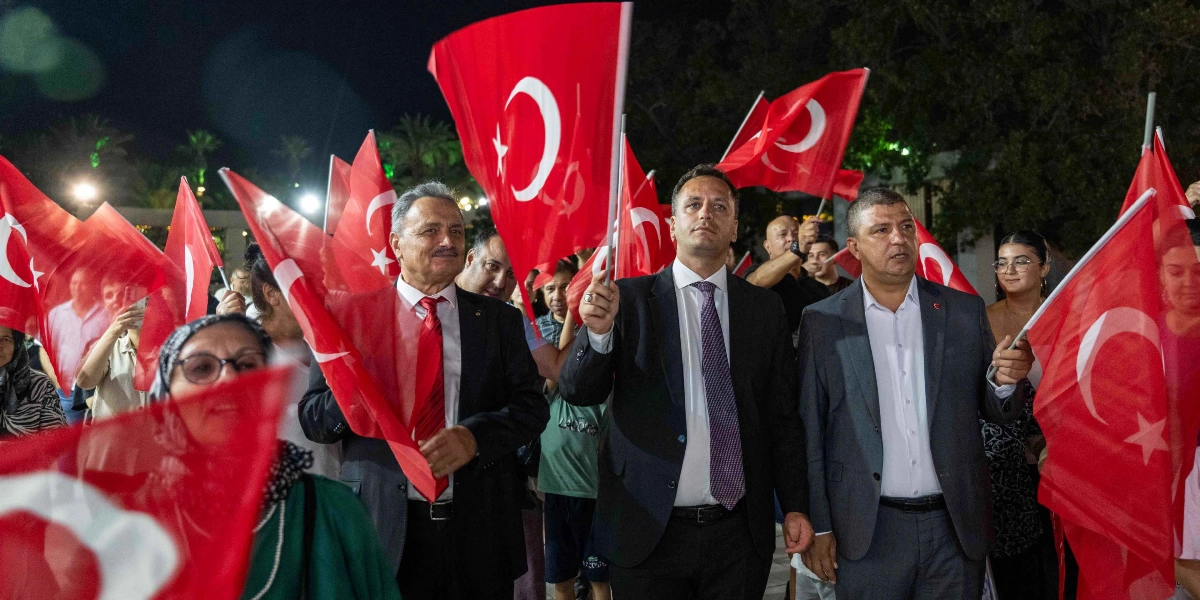 İzmir Enternasyonal Fuarı'nda ellerinde Türk bayrakları tutan bir grup insan, Bornova Belediyesi'ni temsil eden bir etkinlik kapsamında yürüyüş yapıyor. Önde, takım elbiseli üç erkek yan yana ilerlerken, arkalarında ve çevrelerinde bayrak sallayan kalabalık bir grup yer alıyor. Gece vakti dış mekanda çekilen bu karede, katılımcılar coşkulu bir şekilde bayraklarını havaya kaldırıyor.