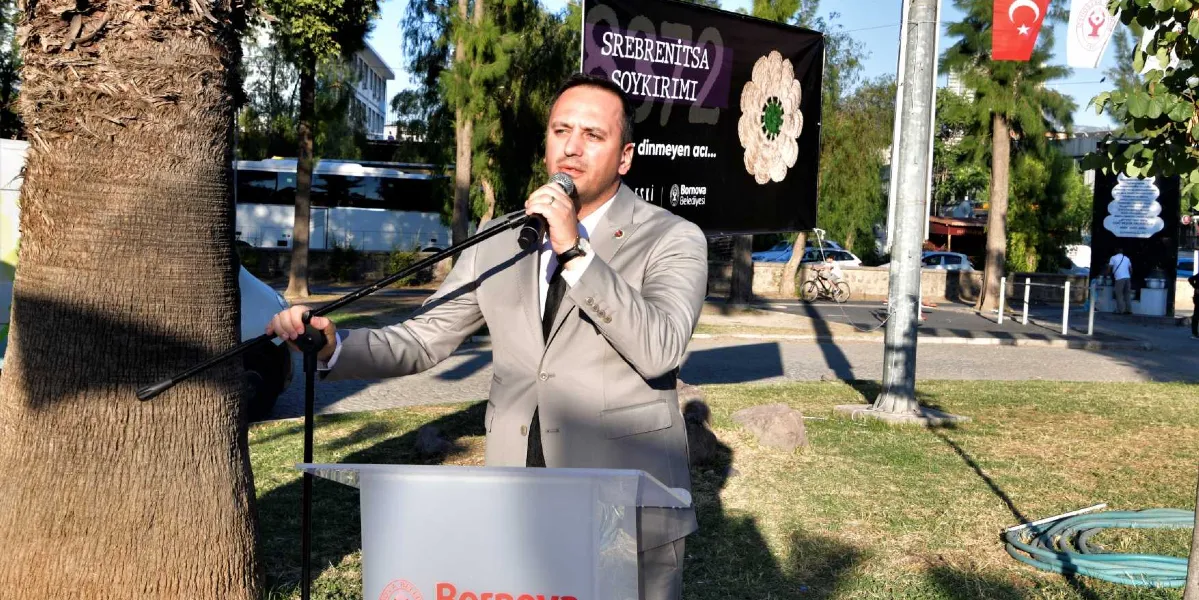 Gri takım elbiseli bir erkek, Bornova Belediyesi logolu bir kürsüde mikrofonla konuşma yapıyor. Arkasında, üzerinde 