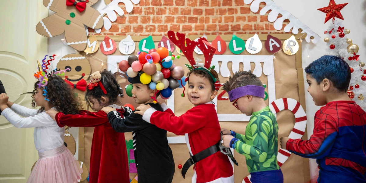 Yeni yıl süslemeleriyle dekore edilmiş bir duvarın önünde birbirlerinin omuzlarından tutarak tren yapmış şekilde yürüyen altı çocuk. Çocuklar çeşitli kostümler ve aksesuarlar giymiş; aralarında başında renkli balonlardan oluşan bir taç olan, geyik boynuzu takan ve Örümcek Adam kostümü giyen çocuklar bulunuyor. Arka planda üzerinde 