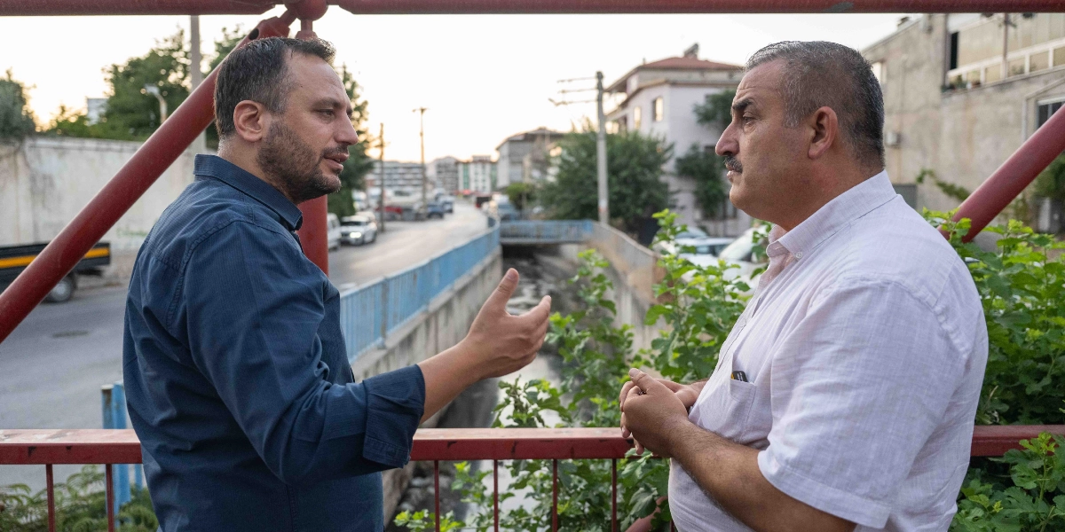Koyu mavi gömlekli bir adam, yanında duran beyaz gömlekli bir adamla bir köprü korkuluğunun önünde sohbet ediyor. Mavi gömlekli adam el hareketleriyle bir şeyler anlatırken, beyaz gömlekli adam onu dinliyor. Arka planda bir dere yatağı, yol ve binalar yer alıyor.