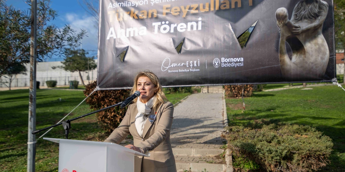 Açık havada, park alanında kurulan bir kürsüde konuşma yapan sarı saçlı bir kadın. Kadının arkasında, üzerinde 
