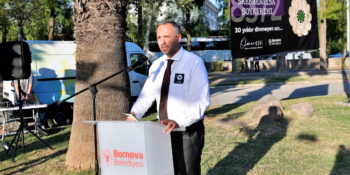 Bornova Belediye Başkanı Ömer Eşki, açık havada kurulan bir kürsüde konuşma yapıyor. Beyaz gömlek ve koyu renk kravat giyen Eşki'nin yakasında Srebrenitsa çiçeği rozeti bulunuyor. Arka planda üzerinde 