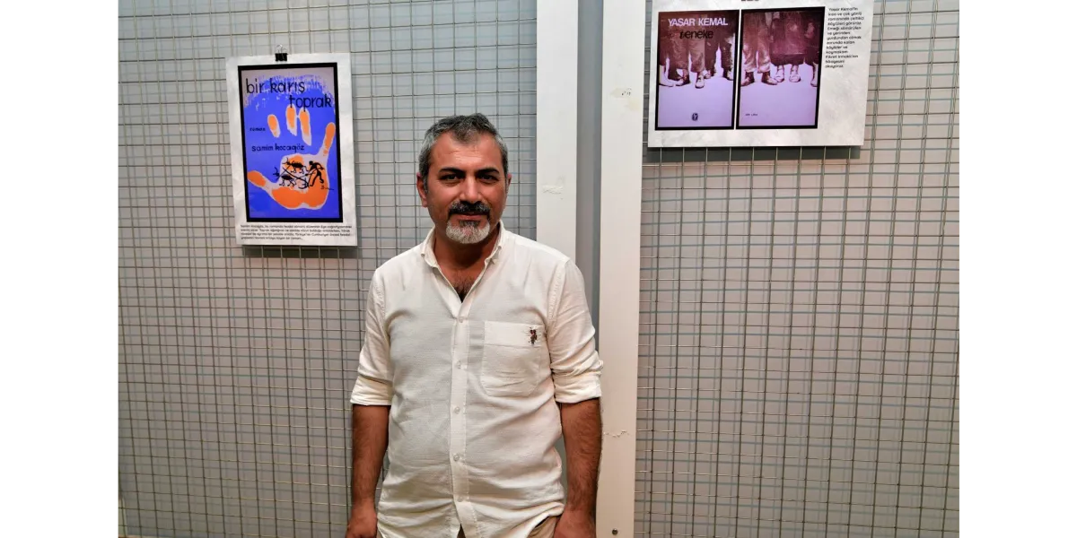Beyaz gömlekli, kirli sakallı bir erkek, arkasında metal bir ızgara panelin bulunduğu sergi alanında duruyor. Panelin sol tarafında Samim Kocagöz’ün 